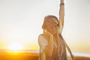 Schöne blonde Frau tanzt beim Hören von Musik über Kopfhörer am Strand gegen den Himmel bei Sonnenuntergang - AJOF00481