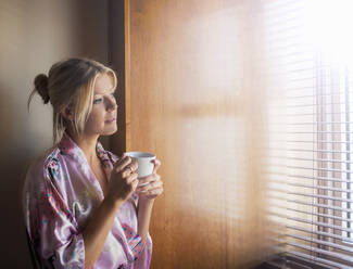 Nachdenkliche junge Frau im Bademantel, die durch das Fenster schaut, während sie zu Hause Kaffee trinkt - AJOF00482