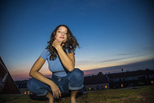 Attractive brunette woman with hand on chin crouching against sky during sunset - NGF00696