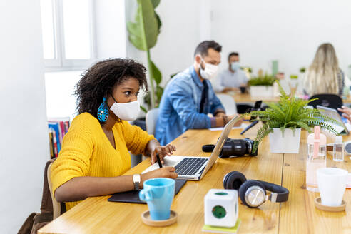 Female wearing face mask working on laptop while sitting with colleague in coworker at office - MPPF01181