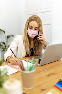 Blond woman wearing face mask writing in book while sitting at office - MPPF01185