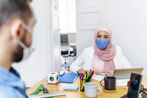 Woman wearing face mask talking with coworker while sitting at office - MPPF01196