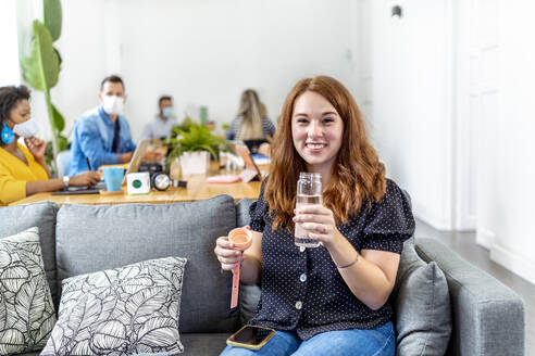 Smiling woman drinking water while sitting with coworker in background at office - MPPF01197