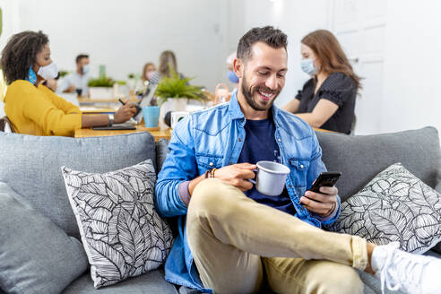 Smiling man holding coffee cup and mobile phone while sitting with coworker in background at office - MPPF01199