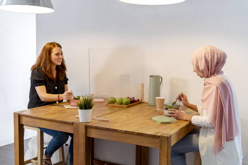 Smiling coworker talking while eating food sitting at screen partition table at office - MPPF01215