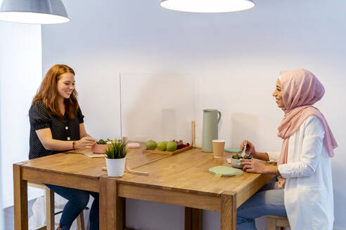 Smiling women having food while sitting at screen partition table in office - MPPF01217