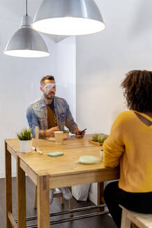 Young woman having food with coworker while sitting at screen partition table in office - MPPF01218