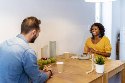 Smiling woman having food with coworker while sitting at screen partition table in office - MPPF01219