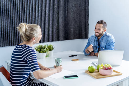 Smiling man and woman drinking coffee while sitting at office - MPPF01231