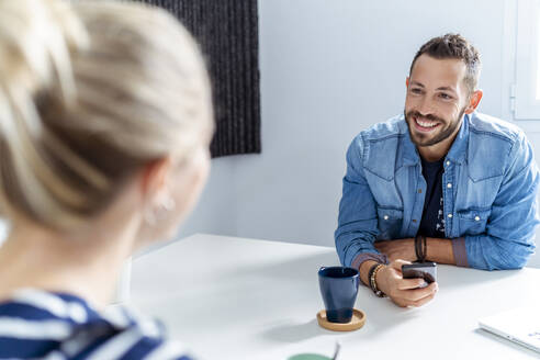 Smiling man talking to woman while having coffee at office - MPPF01232