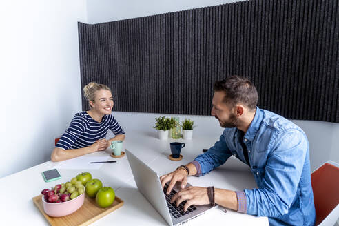 Lächelnder männlicher und weiblicher Mitarbeiter, die im Büro sitzend am Laptop arbeiten - MPPF01235