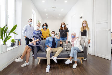 Employees wearing face mask standing and sitting by sofa at office - MPPF01236
