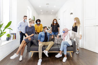 Male and female employee wearing face mask sitting and standing by sofa at office - MPPF01237