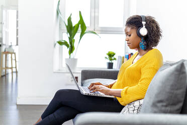 Junge Frau mit Kopfhörern, die einen Laptop benutzt, während sie auf dem Sofa im Büro sitzt - MPPF01243