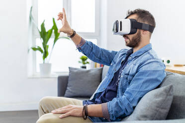 Mid adult man wearing virtual reality simulator working while sitting on sofa - MPPF01245