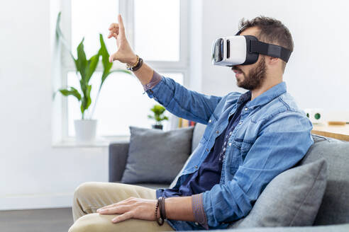 Mittlerer erwachsener Mann, der einen Virtual-Reality-Simulator trägt und auf dem Sofa sitzt - MPPF01245