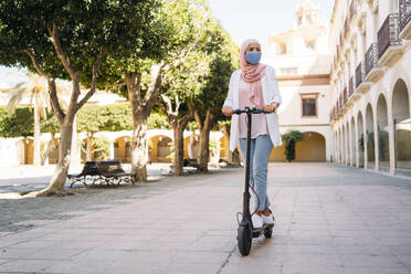 Young woman wearing face mask riding push scooter in city - MPPF01248