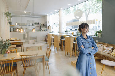 Junge Unternehmerin mit verschränkten Armen, die wegschaut, während sie in einem Café steht - GUSF04550