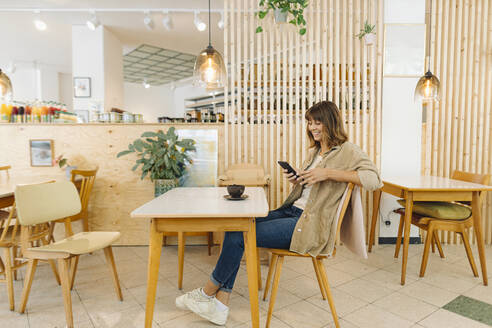 Lächelnde Geschäftsfrau, die in einem Café sitzend eine SMS auf ihrem Smartphone schreibt - GUSF04552