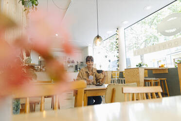 Weibliche Unternehmerin, die eine Textnachricht auf einem Smartphone schreibt und eine Kaffeetasse hält, während sie in einem Café sitzt - GUSF04553