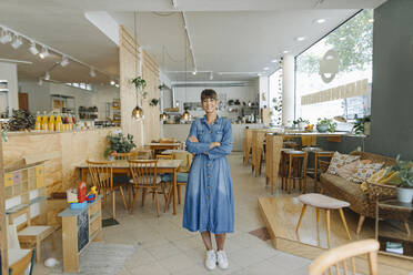 Lächelnde Unternehmerin mit verschränkten Armen in einem Café stehend - GUSF04560