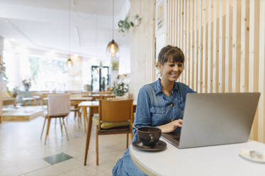 Lächelnde Unternehmerin, die einen Laptop benutzt, während sie in einem Café sitzt - GUSF04563