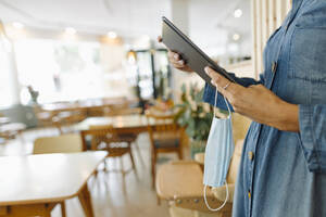 Midsection of female entrepreneur holding digital tablet and protective face mask while standing in cafe - GUSF04566