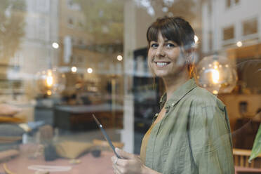 Unternehmerin, die einen digitalen Tisch benutzt und durch ein Fenster schaut, während sie in einem Café steht - GUSF04589