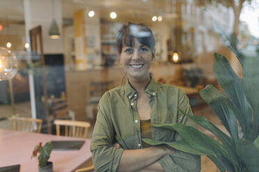 Lächelnde Geschäftsfrau mit verschränkten Armen, die durch das Fenster eines Cafés schaut - GUSF04590