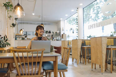 Lächelnde Unternehmerin, die eine Kaffeetasse hält und einen Laptop benutzt, während sie wegschaut und in einem Café sitzt - GUSF04592
