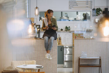 Young businesswoman holding coffee looking away using smart phone sitting on counter in cafe - GUSF04599