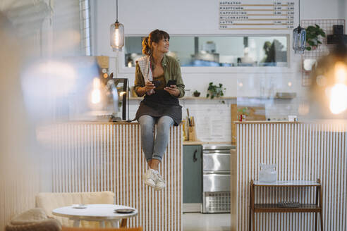 Junge Geschäftsfrau mit Kaffee in der Hand, die wegschaut und ihr Smartphone benutzt, während sie auf dem Tresen eines Cafés sitzt - GUSF04599