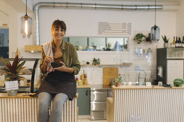 Smiling female owner holding coffee cup sitting on counter in cafe - GUSF04600
