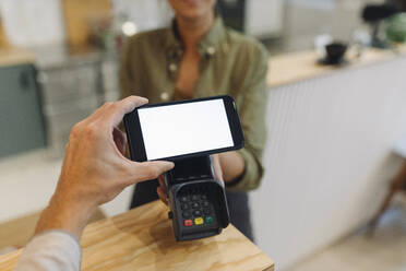 Cropped hand of customer doing contactless payment with smart phone while female owner holding credit card reader at checkout in coffee shop - GUSF04606