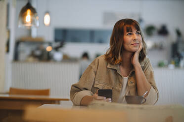 Weibliche Besitzerin mit Hand am Kinn, die ein Smartphone hält, sitzt in einem Café - GUSF04609