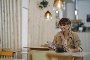 Smiling female owner text messaging on smart phone holding coffee cup sitting in cafe - GUSF04611