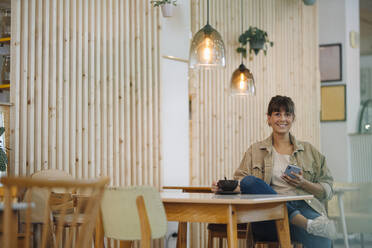 Smiling businesswoman using smart phone while sitting in coffee shop - GUSF04616