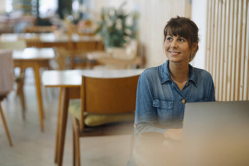 Geschäftsfrau lächelt, während sie in einem Café sitzend ihren Laptop benutzt - GUSF04640