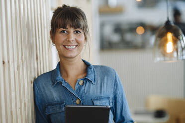 Lächelnde Geschäftsfrau mit digitalem Tablet an einer Holzwand in einem Cafe - GUSF04653