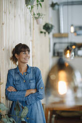 Smiling female entrepreneur looking away with arms crossed by wooden wall - GUSF04654