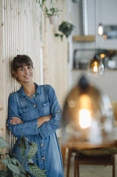 Smiling female owner with arms crossed leaning by wooden wall - GUSF04655