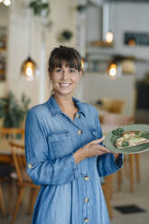Lächelnde Geschäftsfrau, die einen Teller mit Essen hält, während sie in einem Cafe steht - GUSF04659
