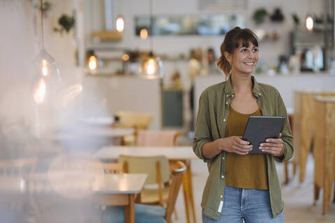 Smiling female entrepreneur looking away while holding digital tablet standing in cafe stock photo