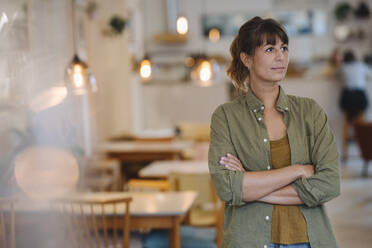 Female entrepreneur with arms crossed looking away while standing in coffee shop - GUSF04679