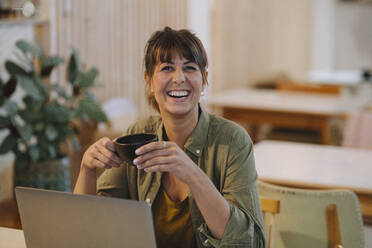 Weibliche Besitzerin lacht, während sie eine Kaffeetasse hält und einen Laptop in einem Café benutzt - GUSF04681