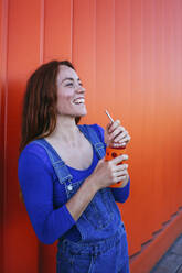 Lächelnde Frau, die einen Saft trinkt, während sie an einer orangefarbenen Wand steht - MGRF00012