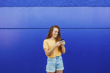 Smiling woman using mobile phone while standing against blue wall - MGRF00015