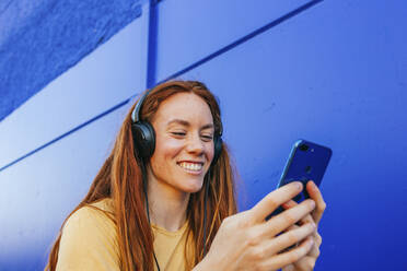 Rothaarige Frau mit Kopfhörern, die ein Mobiltelefon benutzt, während sie an einer blauen Wand sitzt - MGRF00016