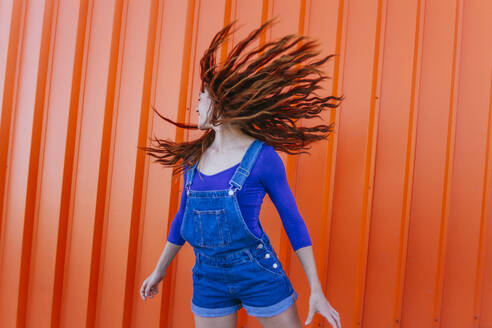 Verspielte Frau, die sich die Haare zuwirft, während sie an einer orangefarbenen Wand steht - MGRF00021