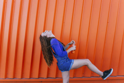Fröhliche Frau trinkt Saft, während sie an einer orangefarbenen Wand steht - MGRF00022